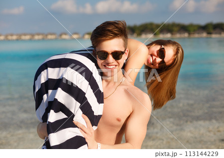 Happy young lovely couple have fun and relax on the beach. Man holds woman on his shoulder. Bungalows of spa resort, palms, and blue lagoon on backgrpund. Family travel concept Happy young lovely couple have fun and relax on the beach. Man holds woman on his shoulder. Bungalows of spa resort, palms, and blue lagoon on backgrpund. Family travel concept 113144229
