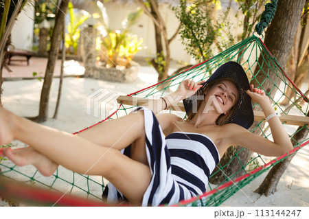 Woman relaxing in the hammock on tropical beach in the shadow, hot sunny day. Girl looks to camera with smile 113144247