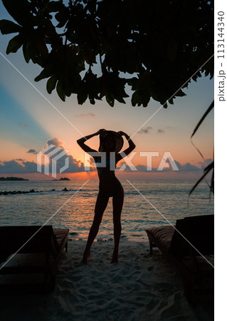 Silhouette of a beautiful slender girl on a sunset background and sun loungers on the ocean shore. Woman watch skyline on sunrise on near deck chair at the sea beach. Vacation Concept. Silhouette of a beautiful slender girl on a sunset background and sun loungers on the ocean shore. Woman watch skyline on sunrise on near deck chair at the sea beach. Vacation Concept. 113144340