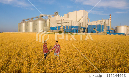 People shaking hands in a wheat field, farmer's agreement. Grain elevator terminal on background. Agriculture agronomist business contract concept. Aerial photo. Agriculture theme 113144468