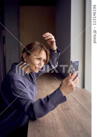 beautiful middle age business woman wearing gray shirt, wide leg pants and black stilettos fix her hair looking into phone camera in modern workspace beautiful middle age business woman wearing gray shirt, wide leg pants and black stilettos fix her hair looking into phone camera in modern workspace 113146198