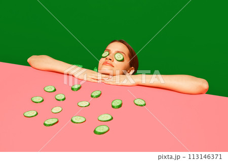 Spa and relaxation. Young woman peeking over table with cucumber slices on face, homemade refreshing mask against green background. Spa and relaxation. Young woman peeking over table with cucumber slices on face, homemade refreshing mask against green background. 113146371