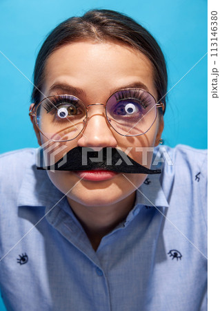 Young woman in shirt, wearing fake moustaches, glasses with eyes items on it and making funny expressions against blue background Young woman in shirt, wearing fake moustaches, glasses with eyes items on it and making funny expressions against blue background 113146380