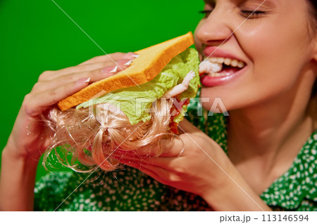 Young woman eating quirky sandwich with doll against red background. Weirdness and surrealism. 113146594