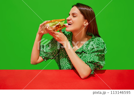 Young woman eating quirky sandwich with doll against red background. Weirdness and surrealism. 113146595