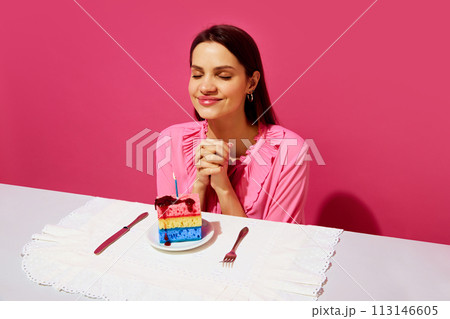 Happy young woman in pink shirt sitting at table with plate and birthday cake made of dishwashing sponges and candle and making birthday wishes Happy young woman in pink shirt sitting at table with plate and birthday cake made of dishwashing sponges and candle and making birthday wishes 113146605