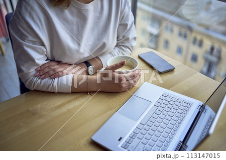 middle age woman with blond hair drinks coffee and works at a laptop in a coworking space with panoramic windows and a view of the city from above, details laptop, coffee, table middle age woman with blond hair drinks coffee and works at a laptop in a coworking space with panoramic windows and a view of the city from above, details laptop, coffee, table 113147015