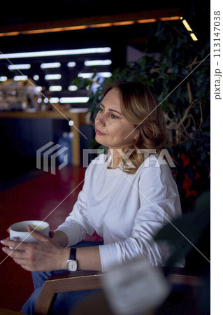 beautiful middle age woman drinking coffee in a beautiful cafe with greenery and light panels in the background 113147038
