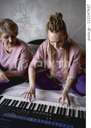 A 60-year-old mother and a 40-year-old daughter play the keyboard together on the bed at home 113147687