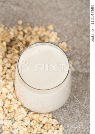 Oat milk in a glass among dry oatmeal flakes on a gray background. 113147699