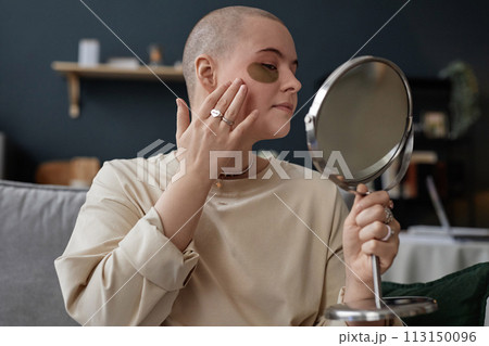 Portrait of young Caucasian woman with undereye patches on face holding mirror applying nourishing cream on skin Portrait of young Caucasian woman with undereye patches on face holding mirror applying nourishing cream on skin 113150096