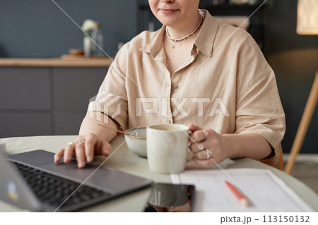 Medium crop shot of young Caucasian woman sitting at table in kitchen having breakfast and scrolling web page in Internet on laptop Medium crop shot of young Caucasian woman sitting at table in kitchen having breakfast and scrolling web page in Internet on laptop 113150132