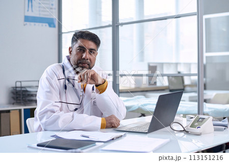 Portrait of senior Middle Eastern doctor sitting at workplace in medical clinic and looking at camera with confidence copy space 113151216