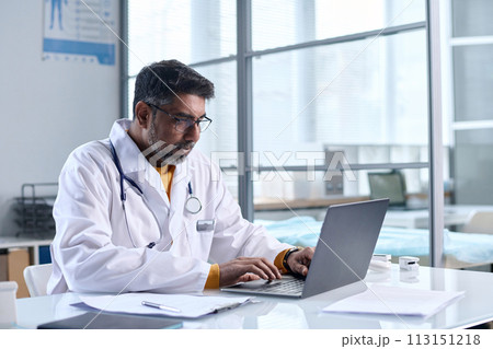 Portrait of senior Middle Eastern doctor using laptop while sitting at desk in medical clinic copy space Portrait of senior Middle Eastern doctor using laptop while sitting at desk in medical clinic copy space 113151218
