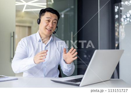Professional Asian businessman in a headset engaged in a video call, explaining with hand gestures in a bright office. Professional Asian businessman in a headset engaged in a video call, explaining with hand gestures in a bright office. 113151727