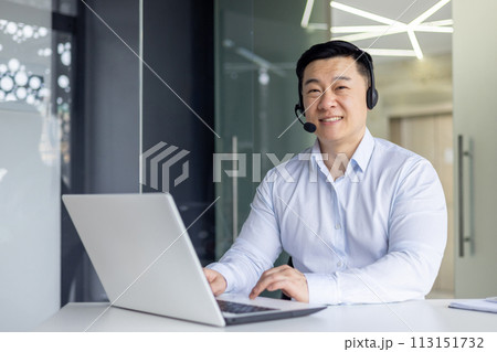 A cheerful Asian man with a headset is working on a laptop in a contemporary office setting, radiating professionalism and positivity. A cheerful Asian man with a headset is working on a laptop in a contemporary office setting, radiating professionalism and positivity. 113151732