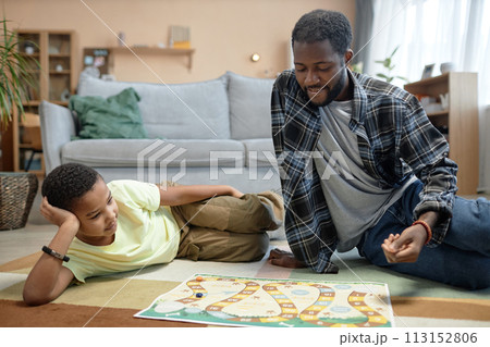 Full length portrait of Black father and son playing board game together sitting on floor at home enjoying family time 113152806