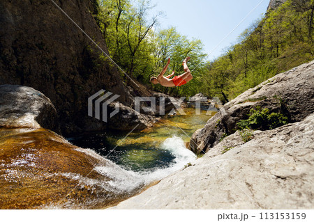 Young Man Jumping From Cliff Into Water of Mountain River 113153159
