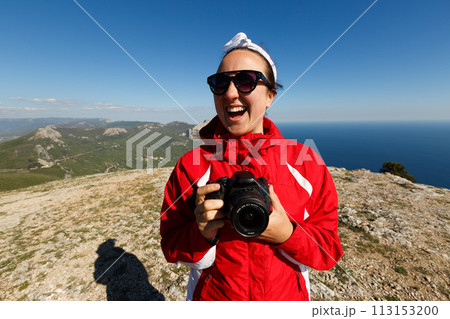 Happy woman photographer smiles on a peak of a mountain with dslr camera in her hands in mountains enjoying the climb with beautiful rocky and sea background 113153200