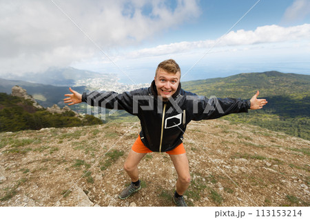 Happy smiling man stands on a top of the mountain with open hands and strong expession looking at camera with summer mountain landscape on hiking trip 113153214