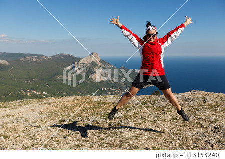 Happy woman jumps on a peak of a mountain in summer time in mountains enjoying the climb with beautiful rocky and sea background 113153240