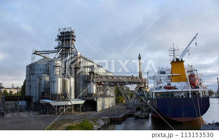 Big grain terminal at seaport. Cereals bulk transshipment from road transport to vessel. Loading grain crops on ship from large elevators at the berth. Transrportation of agricultural products. Big grain terminal at seaport. Cereals bulk transshipment from road transport to vessel. Loading grain crops on ship from large elevators at the berth. Transrportation of agricultural products. 113153530