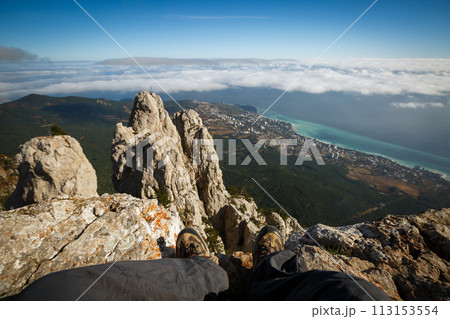 Traveler sitting on a cliff of rocky mountain summit above clouds. Point of view photo of legs in hiking boots with sea, city and mountain landscape. Ai Petri, Yalta, Crimea. Traveler sitting on a cliff of rocky mountain summit above clouds. Point of view photo of legs in hiking boots with sea, city and mountain landscape. Ai Petri, Yalta, Crimea. 113153554