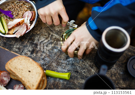 Hiker opens cans on wooden table during breakfast at the forest camp. People camping at national park and doing lunch. Isolated view 113153561