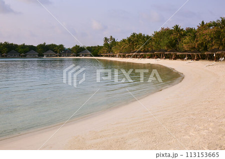 Indian ocean coastline on Maldives island. Beach chairs under straw umbrellas. White sandy beach and calm sea. Bungalows on background 113153665
