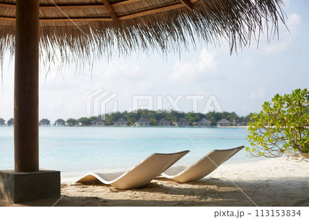 Two modern beach chairs on the beach under straw umbrella. Indian ocean coastline on Maldives island. White sandy shore and calm sea. Travel and vacation concept. 113153834