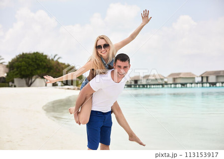 Portrait of smiling man carrying woman on his back along the sea shore. Guy giving piggyback ride to girlfriend by ocean. Young couple having fun on tropical resort beach. Honeymoon concept. 113153917