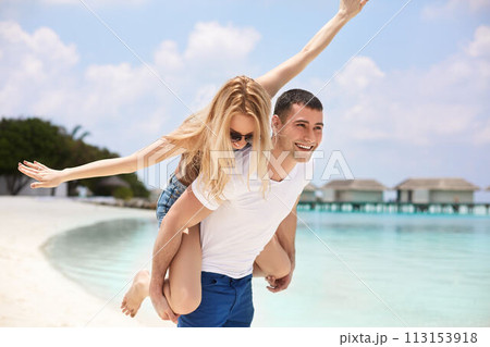 Portrait of smiling man carrying woman on his back along the sea shore. Guy giving piggyback ride to girlfriend by ocean. Young couple having fun on tropical resort beach. Honeymoon concept. 113153918