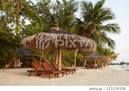 Beach deck chairs under straw umbrellas. Indian ocean coastline on Maldives island. White sandy beach and calm sea. Travel and vacation concept. Beach deck chairs under straw umbrellas. Indian ocean coastline on Maldives island. White sandy beach and calm sea. Travel and vacation concept. 113154108
