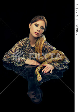 Portrait of young woman with makeup, in animal print blouse posing with snake against black studio background. Deep feelings Portrait of young woman with makeup, in animal print blouse posing with snake against black studio background. Deep feelings 113155185
