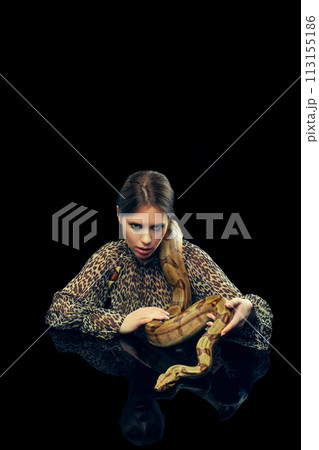 Portrait of young woman with makeup, in animal print blouse posing with snake against black studio background. Portrait of young woman with makeup, in animal print blouse posing with snake against black studio background. 113155186