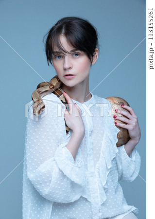 Portrait of tender young woman wit blue eyes, nude makeup, in white dress posing with snake against grey-blue studio background. 113155261