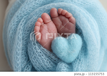 The tiny foot of a newborn baby. Soft feet of a new born in a blue wool blanket. Close up of toes, heels and feet of a newborn. Knitted blue heart in the legs of a baby. Macro photography.  113155753