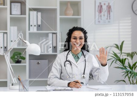 Friendly Latina doctor in a white coat greeting a patient during a virtual consultation in her office. 113157058