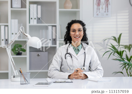 Professional Hispanic female doctor sitting at her desk with a stethoscope, ready to start a video call in a clinic office setting. Professional Hispanic female doctor sitting at her desk with a stethoscope, ready to start a video call in a clinic office setting. 113157059