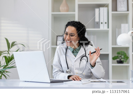 A cheerful hispanic female doctor is seen talking to a patient during a video call consultation from her office. A cheerful hispanic female doctor is seen talking to a patient during a video call consultation from her office. 113157061