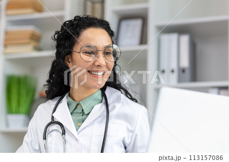 A joyful Latina doctor wearing glasses smiles cheerfully at work, radiating positivity and confidence in a clinical setting. 113157086
