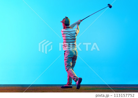 Back view portrait of young man, professional golfer playing golf on green lash grass in neon light against gradient blue background. 113157752