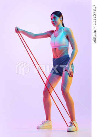 Side view of focused, brunette woman exercising with resistance band in neon light against gradient studio background. 113157817