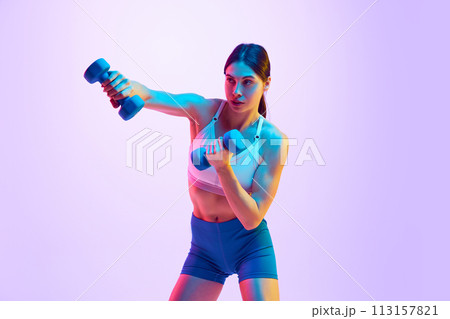 Young attractive woman in activewear boxing with dumbbells in hand in motion in neon light against gradient studio background. 113157821