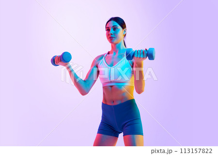 Young athlete woman doing exercises with dumbbells for biceps in neon light against gradient studio background. Young athlete woman doing exercises with dumbbells for biceps in neon light against gradient studio background. 113157822