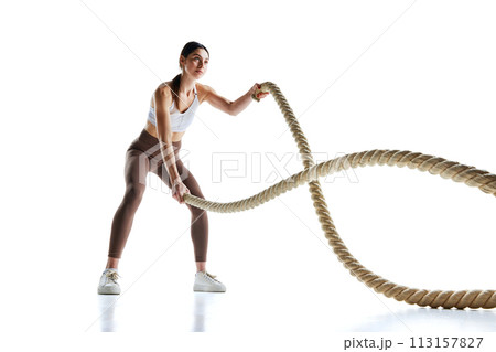 Young strong fit woman doing exercises with battle ropes at gym against white studio background. 113157827