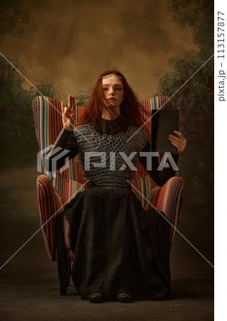 Young woman, dressed as brave knight sitting on chair and holds modern tablet with blank screen against vintage studio background. 113157877