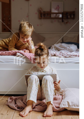 Siblings watching something on tablet, boy lying on bed, cute girl sitting on on floor in bedroom. Children's screen time. 113157908