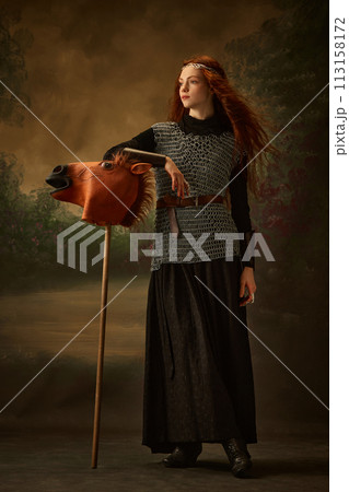 Red-haired princess in chainmail and black dress touching whimsical horse head on stick, toy against vintage studio background. Red-haired princess in chainmail and black dress touching whimsical horse head on stick, toy against vintage studio background. 113158172