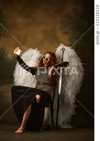 Redhaired angel in warrior chainmail with sword takes selfie on modern smartphone against vintage studio background. 113158210